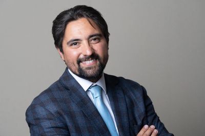 Studio headshot of Pete Gonzales in a navy checked suit and light blue tie, arms folded.