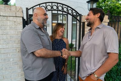 Pete Gonzales shaking hands with a resident at a front gate while a neighbor laughs beside them.